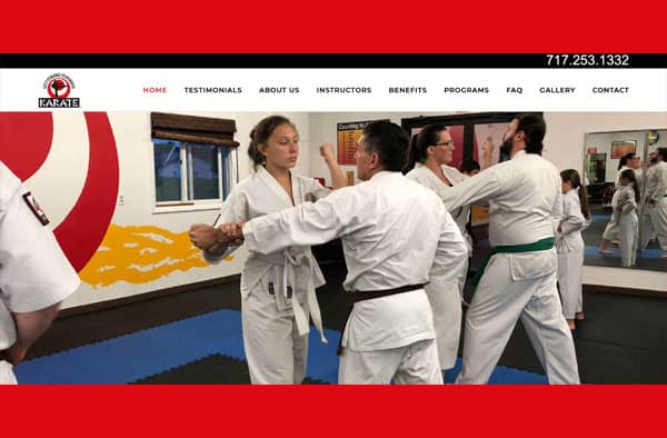 A group of people in white karate uniforms practice self-defense moves in a dojo, as one instructor guides a student. Mats cover the floor, and a large red and white wall logo—perfect for a website portfolio—stands out in the background.